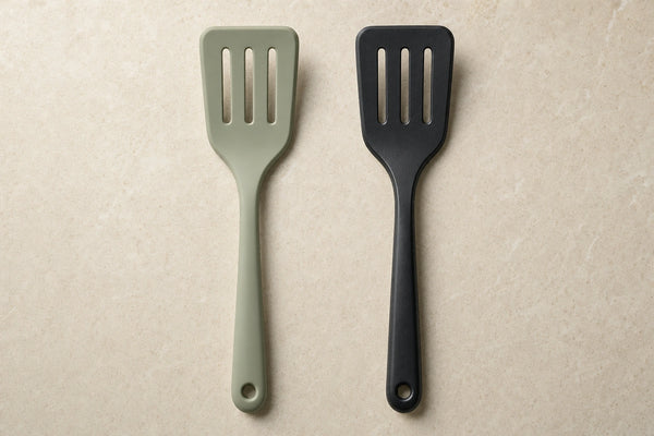 Illustrative side-by-side comparison of a muted sage silicone slotted turner and a black plastic slotted turner on a pale stone surface.