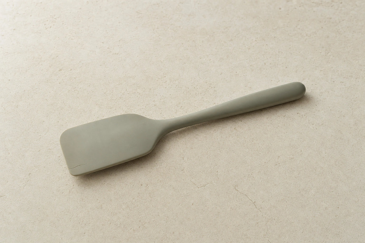 Muted sage silicone spatula with a small split along one edge on a pale stone surface.
